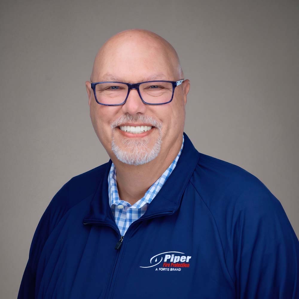 Smiling bald man with glasses and goatee wearing a blue Piper Fire Protection jacket over a blue and white checkered shirt against a neutral gray background.