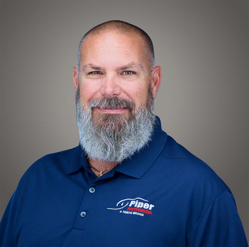 Portrait of a bald man with a thick gray beard wearing a navy blue Polo shirt with a 'Piper Fire Protection A Fortis Brand' logo on the chest, against a neutral gray background.