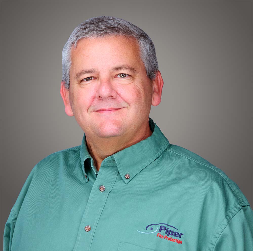Smiling middle-aged man with short gray hair wearing a green button-up shirt with a Piper Fire Protection logo on the chest, against a neutral gray background.