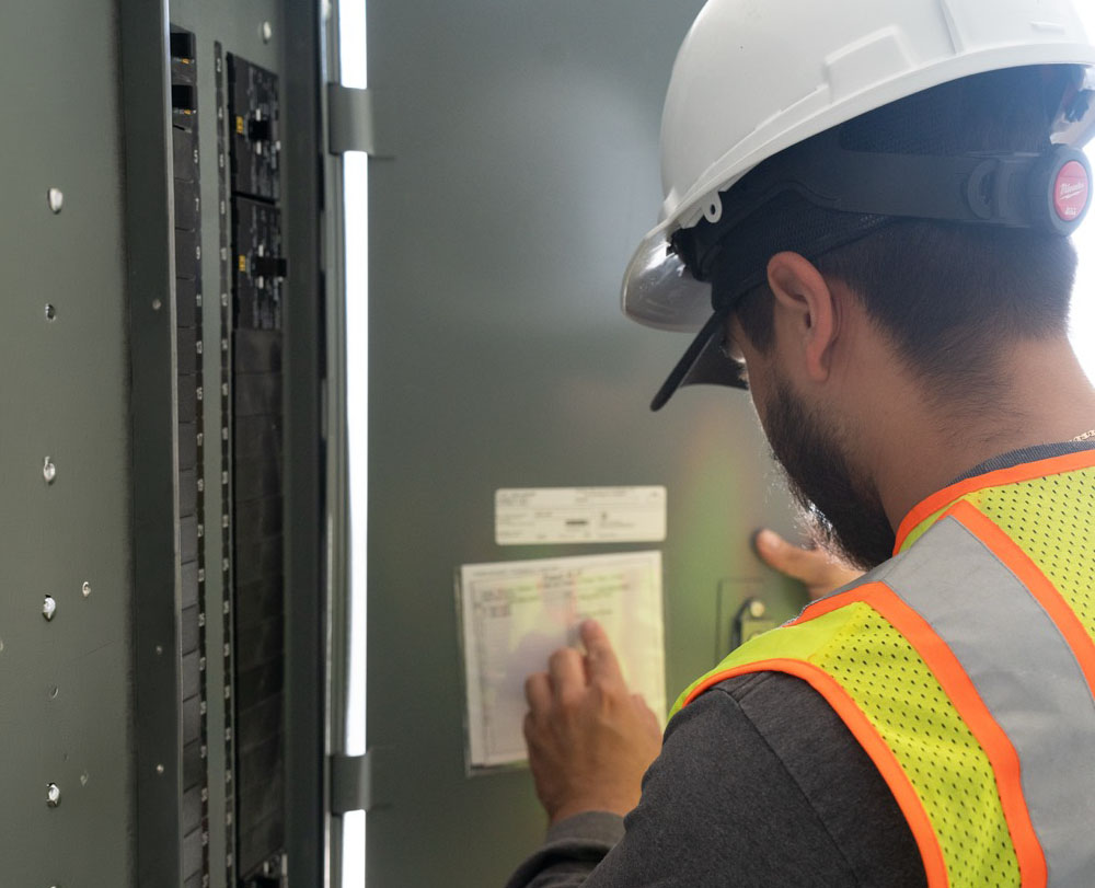 Man wearing a white hard hat and high-visibility safety vest examining the labels on an electrical panel inside a grey metal enclosure.