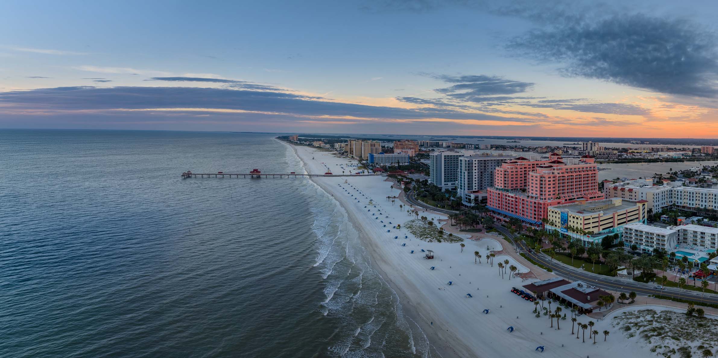 Aerial view of a long sandy beach in Clearwater, FL with blue umbrellas and palm trees next to a coastal city with tall buildings at sunset, featuring a pier extending into calm ocean waters under a partly cloudy sky.