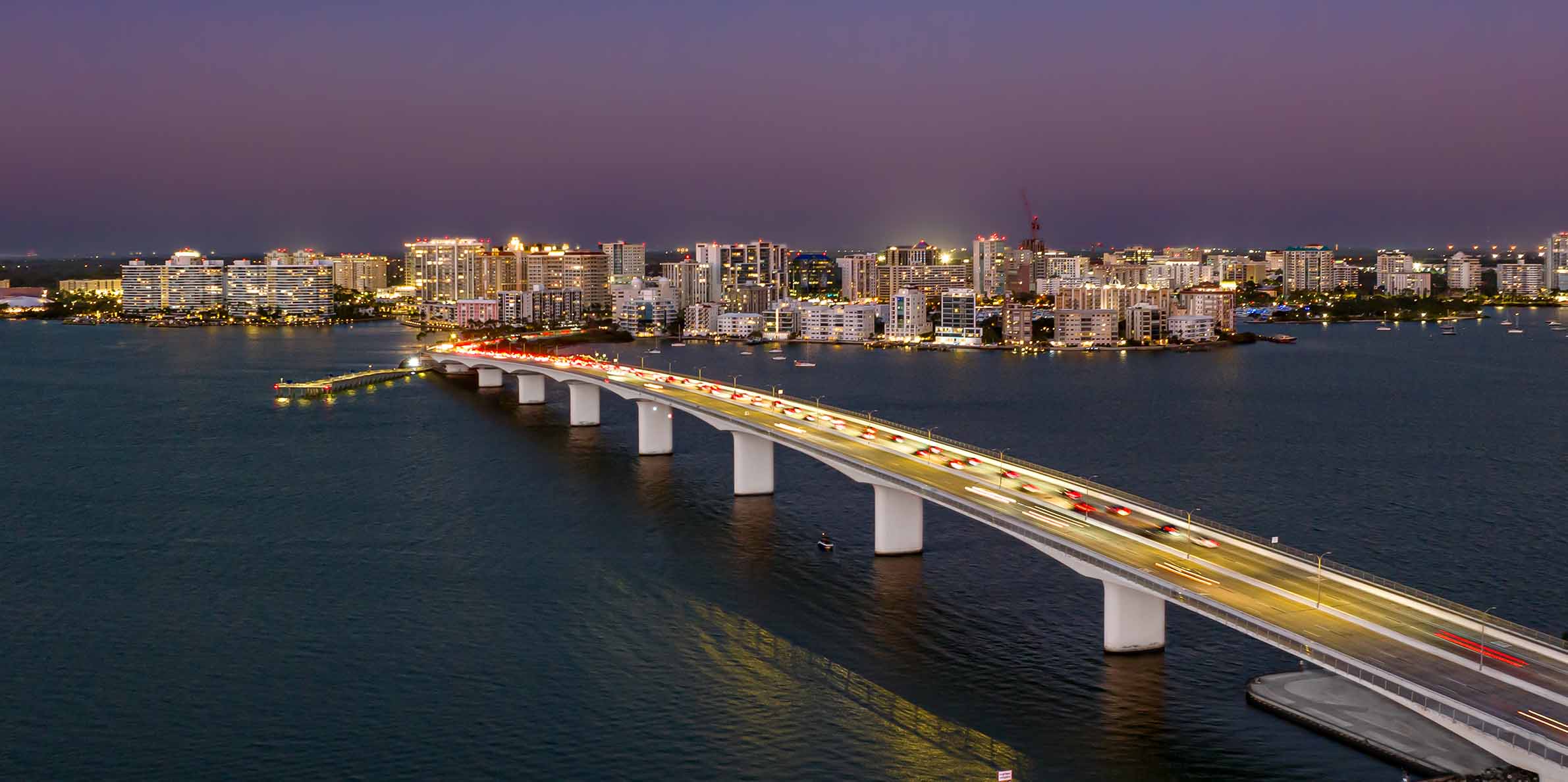 A long bridge in Sarasota, FL lit with vehicle lights stretches across water toward a brightly illuminated city skyline at dusk.