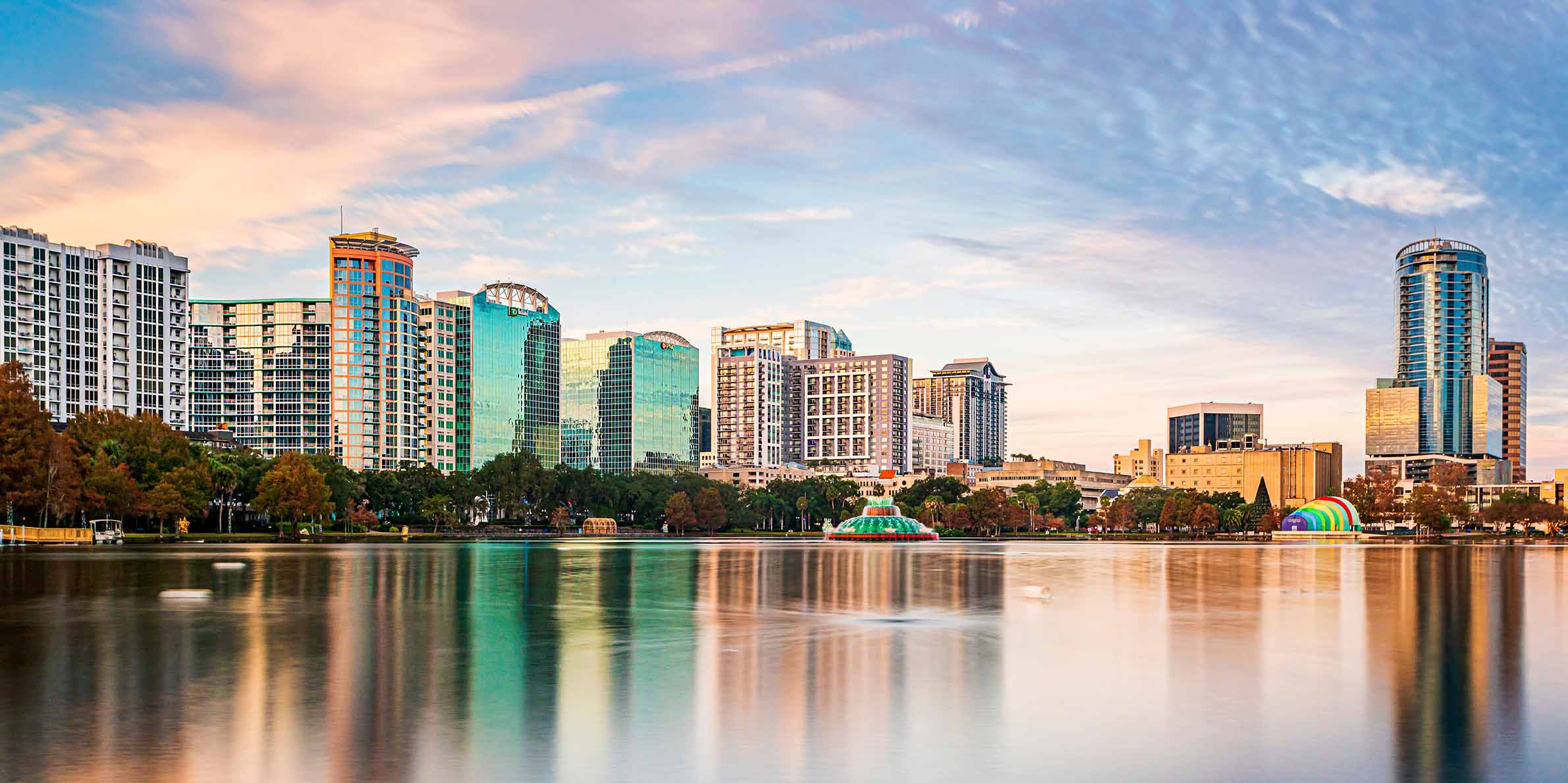 Orlando city skyline at sunset with tall modern buildings reflecting on a calm lake, framed by trees along the shoreline.