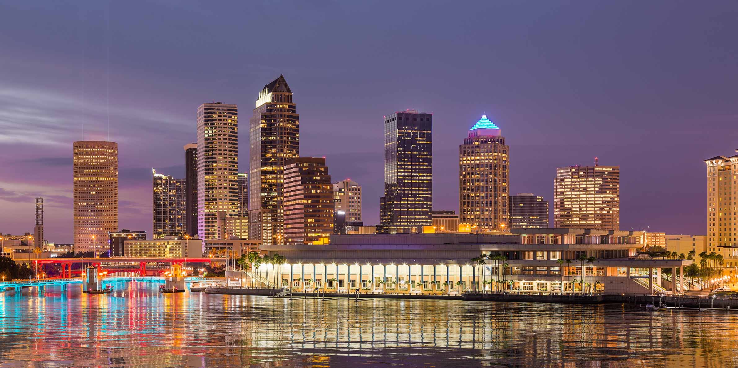 Tampa skyline at dusk with illuminated skyscrapers and their reflections on calm water, including a building with a blue-lit pyramid roof and a red-lit bridge.