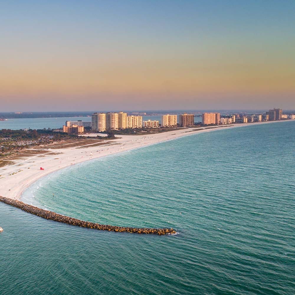Aerial view of a curved white sandy beach in Clearwater, FL along a coastal city with multiple high-rise buildings, calm turquoise water, and a rocky pier extending into the ocean at sunset.