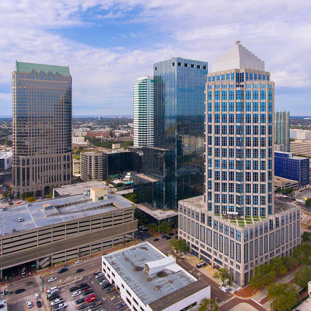 Aerial view of downtown Tampa with several tall office buildings under a partly cloudy sky, surrounded by streets with cars and smaller buildings.