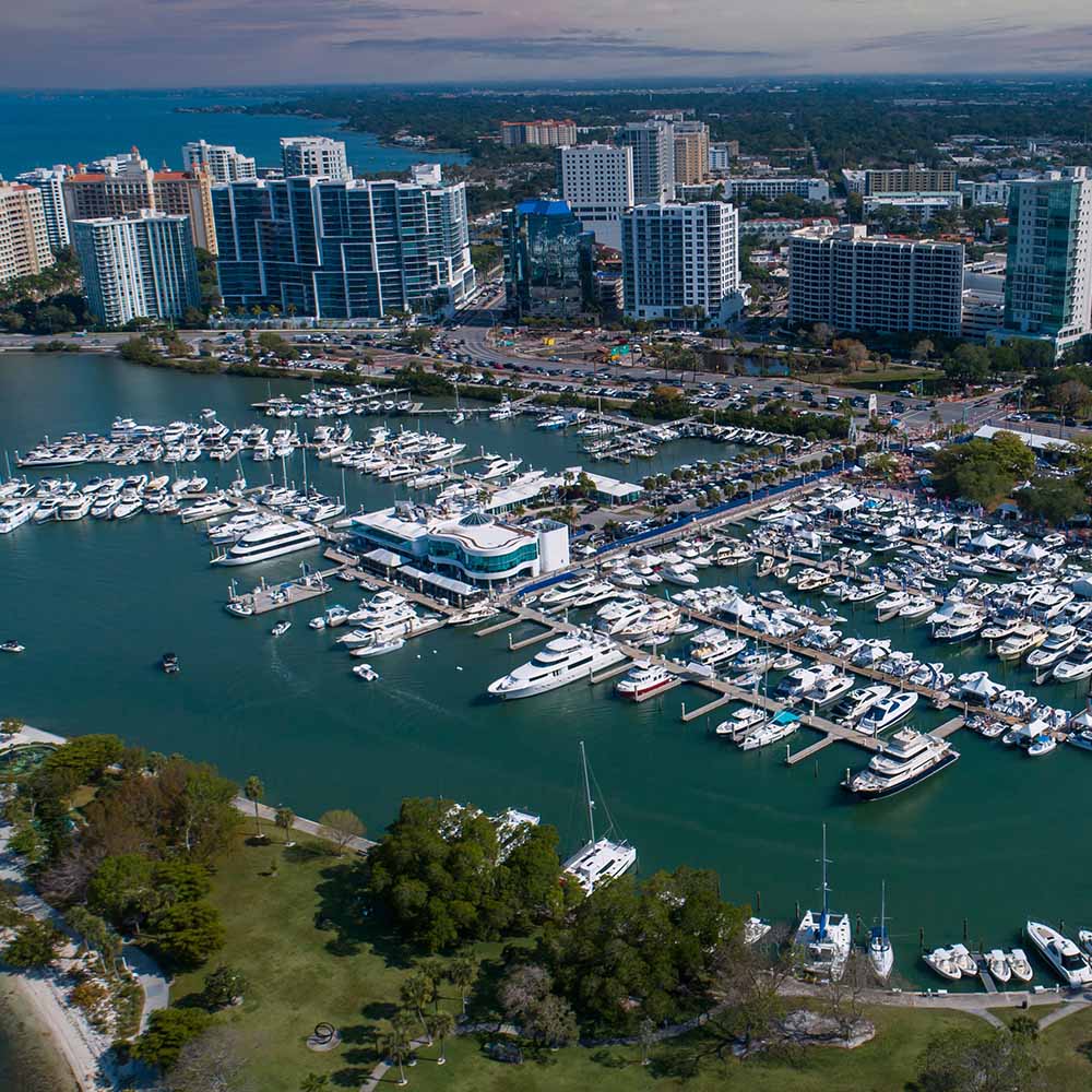 Aerial view of a Sarasota marina filled with many yachts and boats docked in calm blue-green water, with a modern white building near the center, surrounded by a city skyline of mid-rise buildings and greenery under a partly cloudy sky.