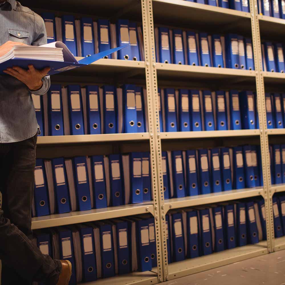 A person in a gray shirt holding and reviewing documents in a blue folder, standing next to shelves filled with neatly arranged blue binders.