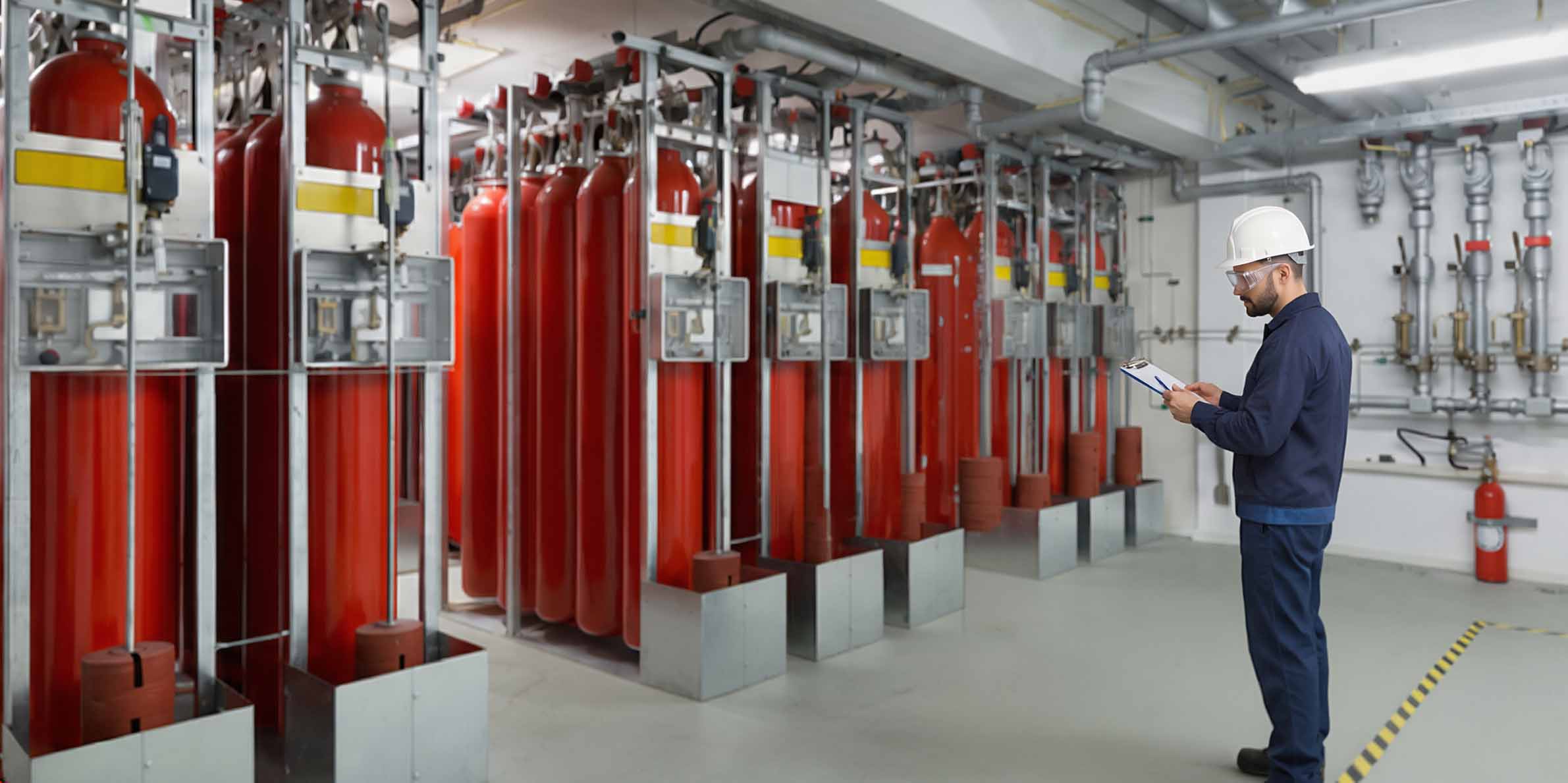 Man in safety helmet and goggles holding clipboard and inspecting large red industrial gas cylinders in a clean facility with metal piping on the wall.