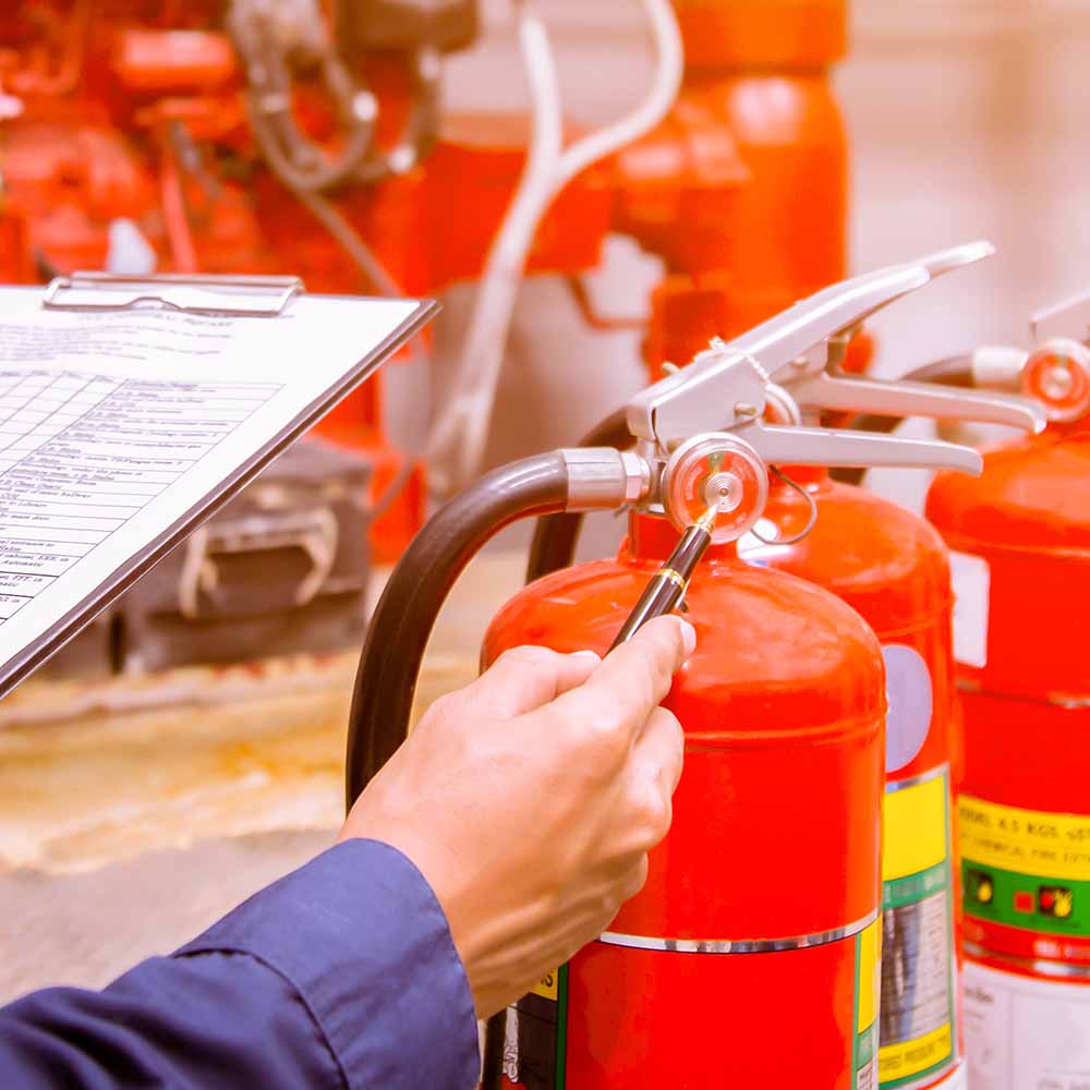 Person inspecting the pressure gauge of a red fire extinguisher, with more fire extinguishers and a clipboard with a checklist in the background.