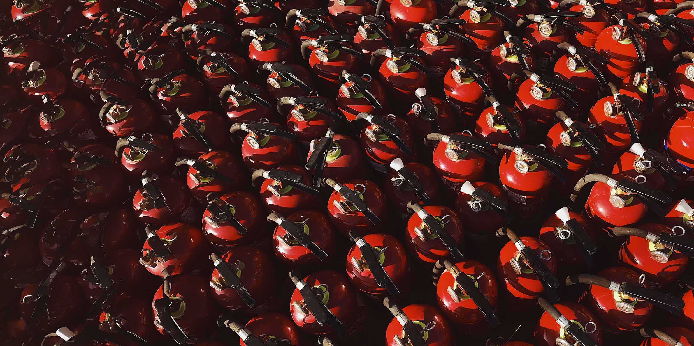 Many red fire extinguishers densely arranged in rows, with black handles and pressure gauges visible on top.