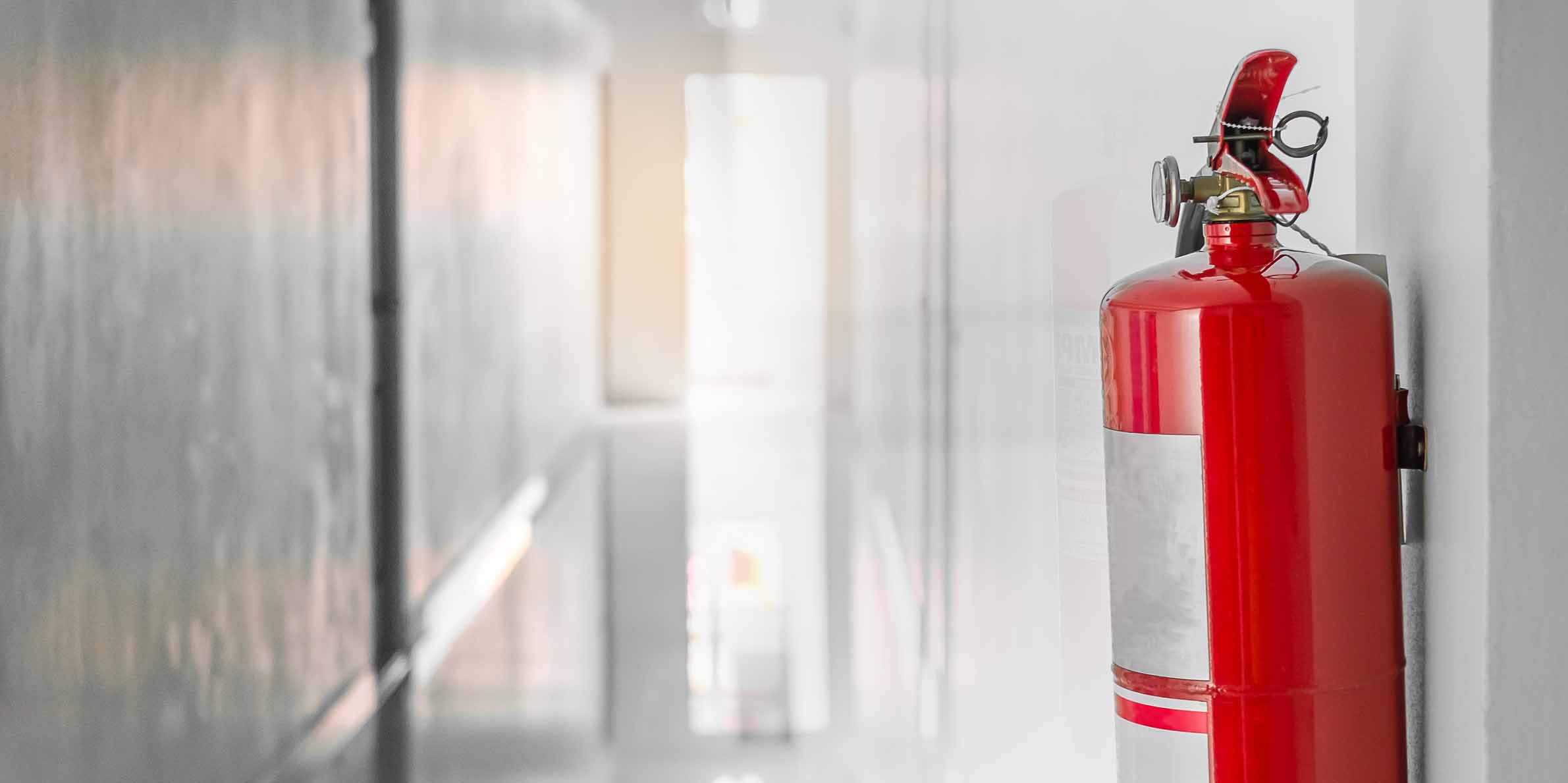 Red fire extinguisher mounted on a white wall in a well-lit corridor with a blurry background.
