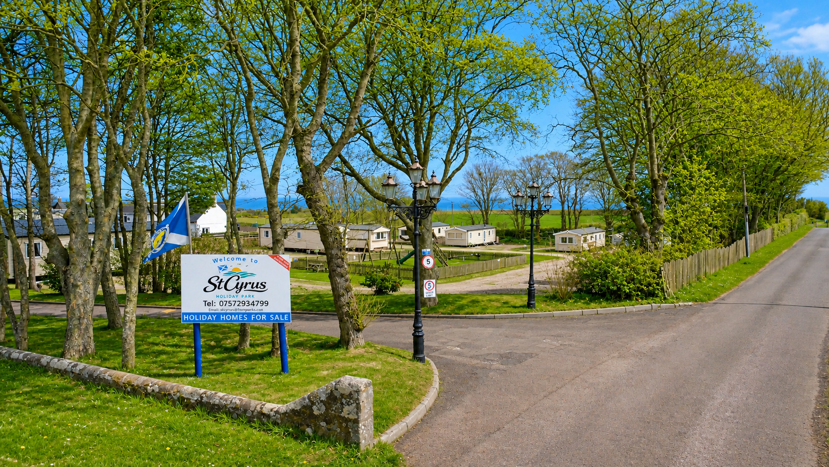 St Cyrus holiday park entrance sign with tree-lined road, caravans, and coastal landscape.