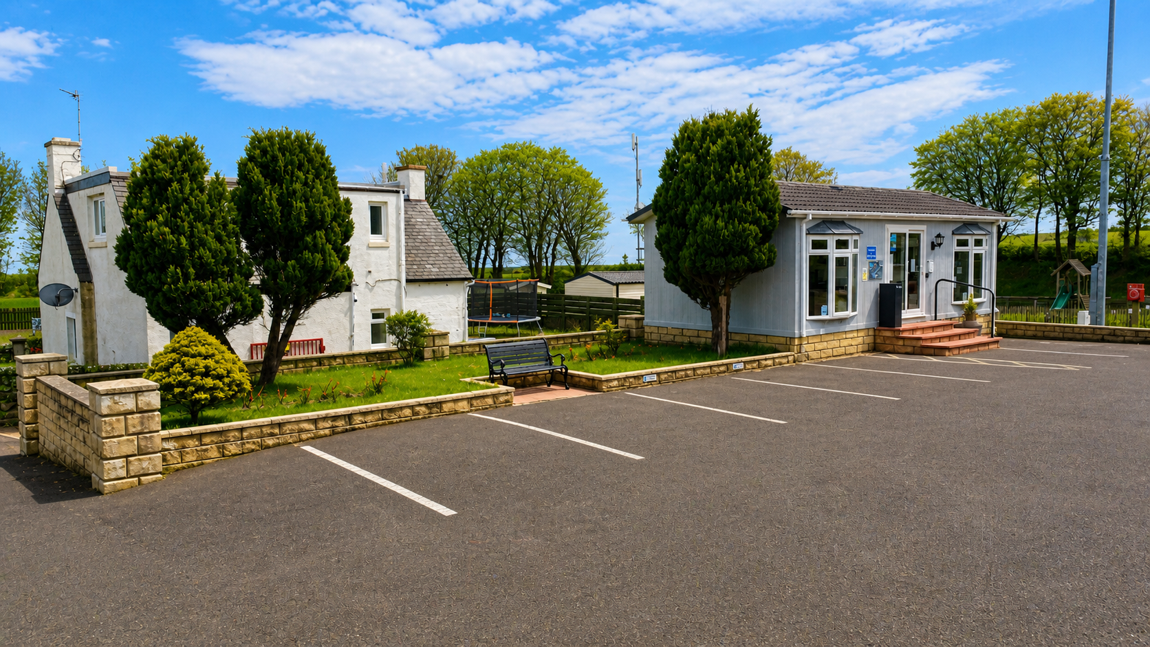 Rural Scottish buildings with hedges, green lawn, and paved parking area under blue sky