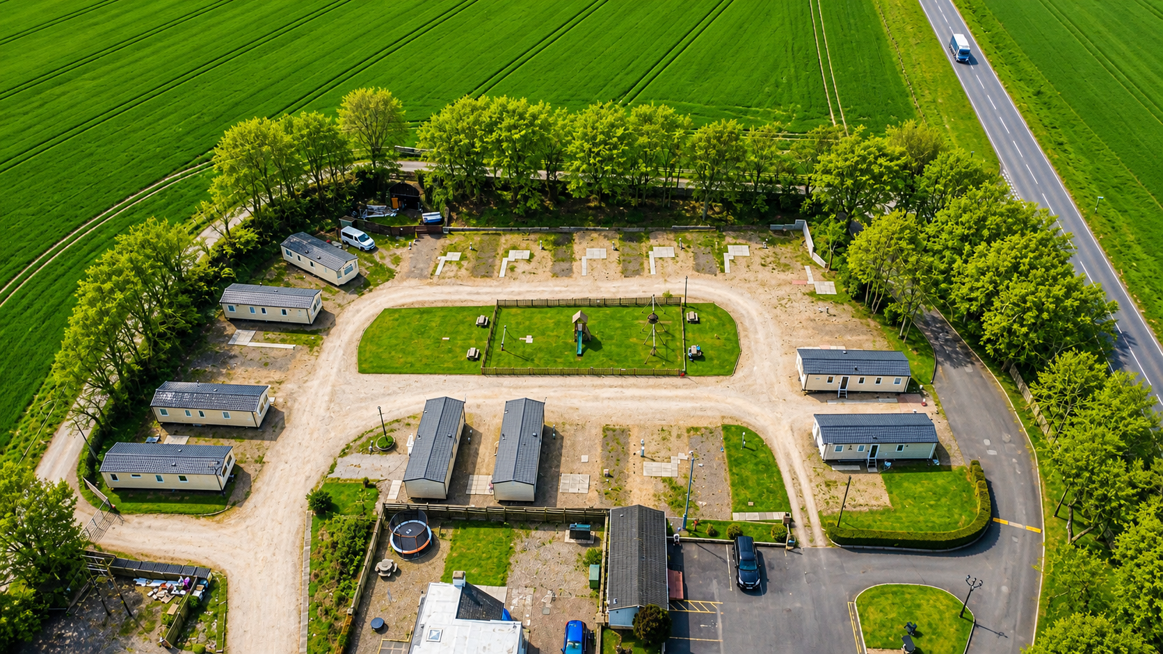 Aerial view of rural campground with mobile homes, green fields, trees, and paved road.