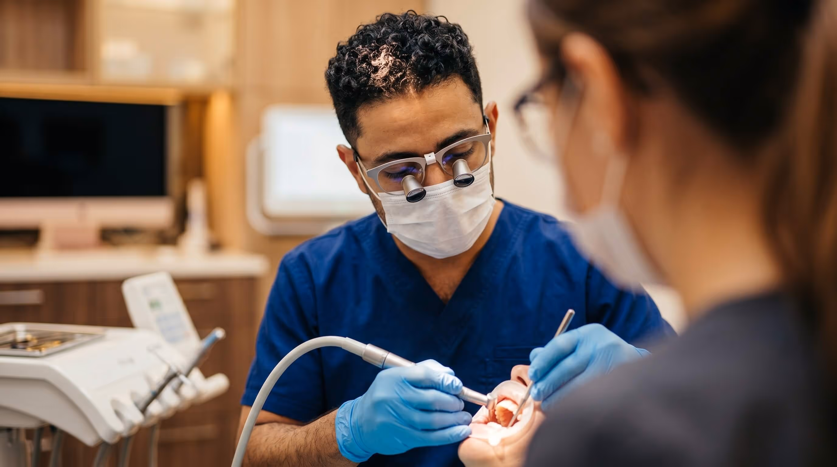 Dentist in blue scrubs and magnifying glasses performing dental treatment on a patient.