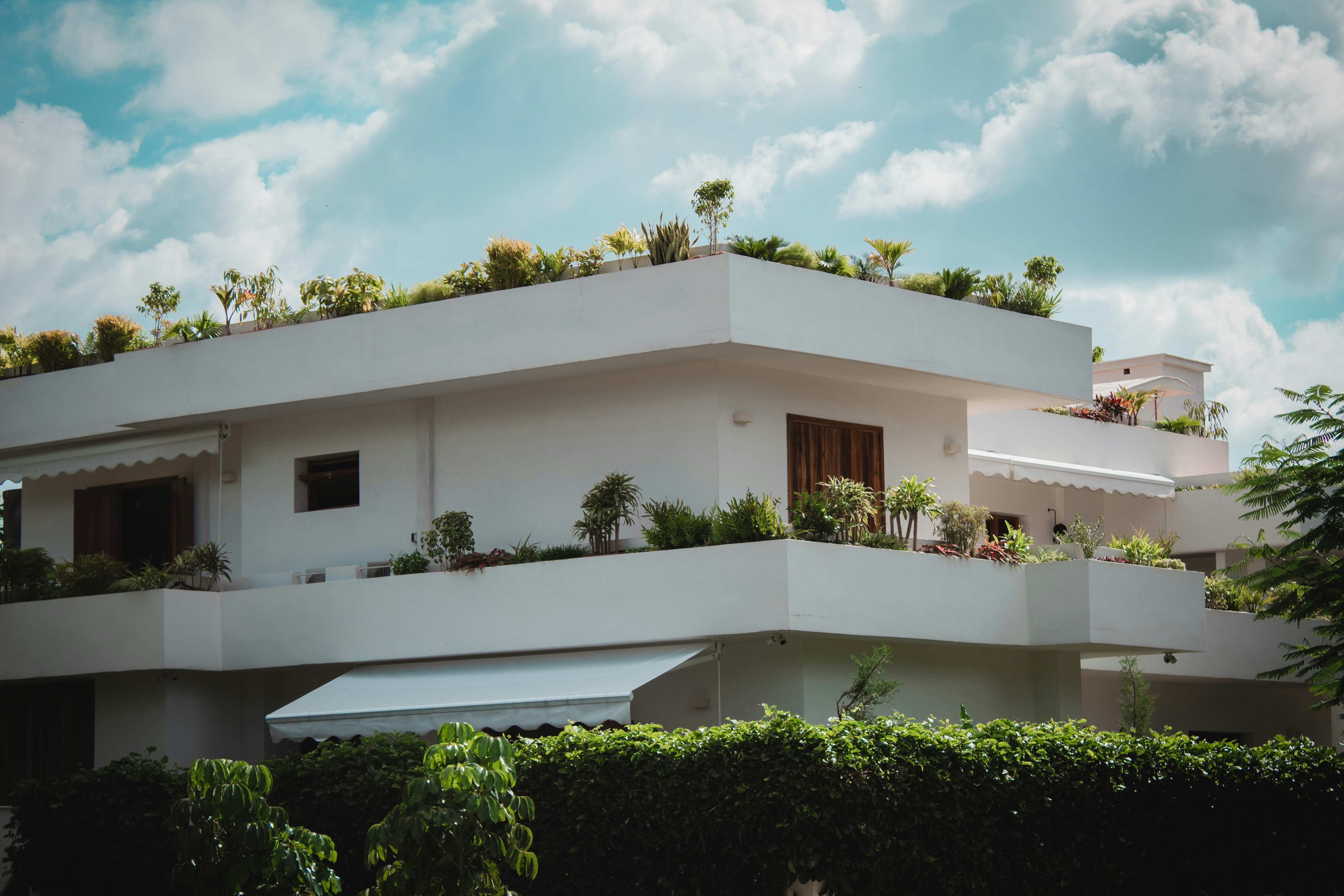 Modern white building with plants on balconies and rooftop under a partly cloudy sky.