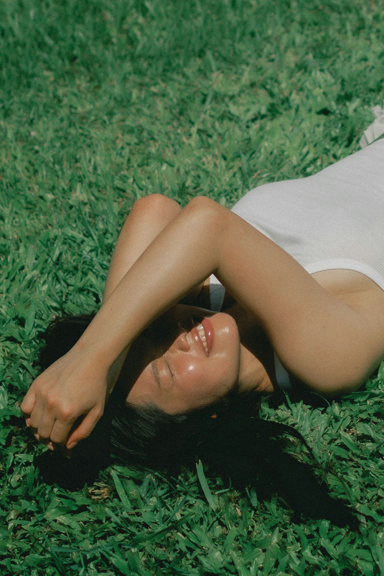 Woman lying on green grass with eyes closed and smiling, shielding face from sunlight with her arm.