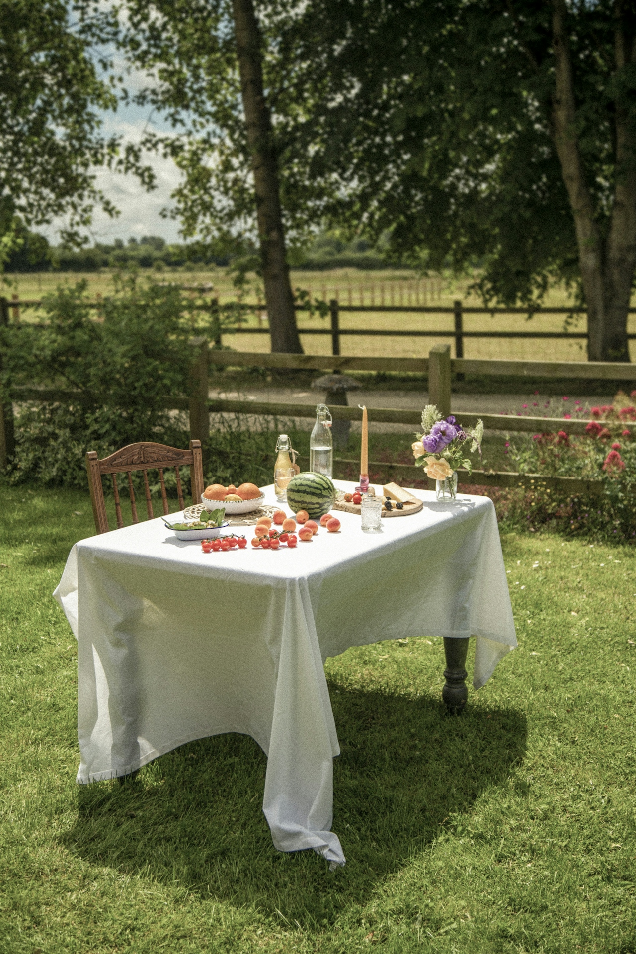 Outdoor table with a white tablecloth displaying a watermelon, apricots, cherry tomatoes, bottles of water, a candle, and a vase of flowers on green grass under trees.