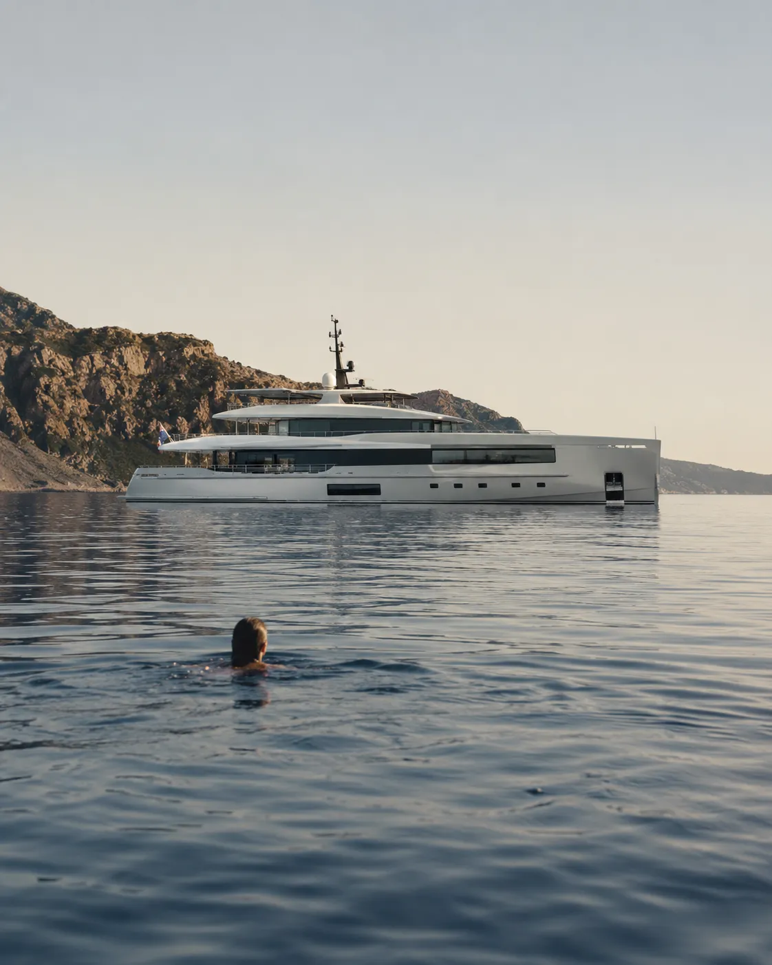 Person swimming in calm sea near a large modern yacht with rocky cliffs in the background.