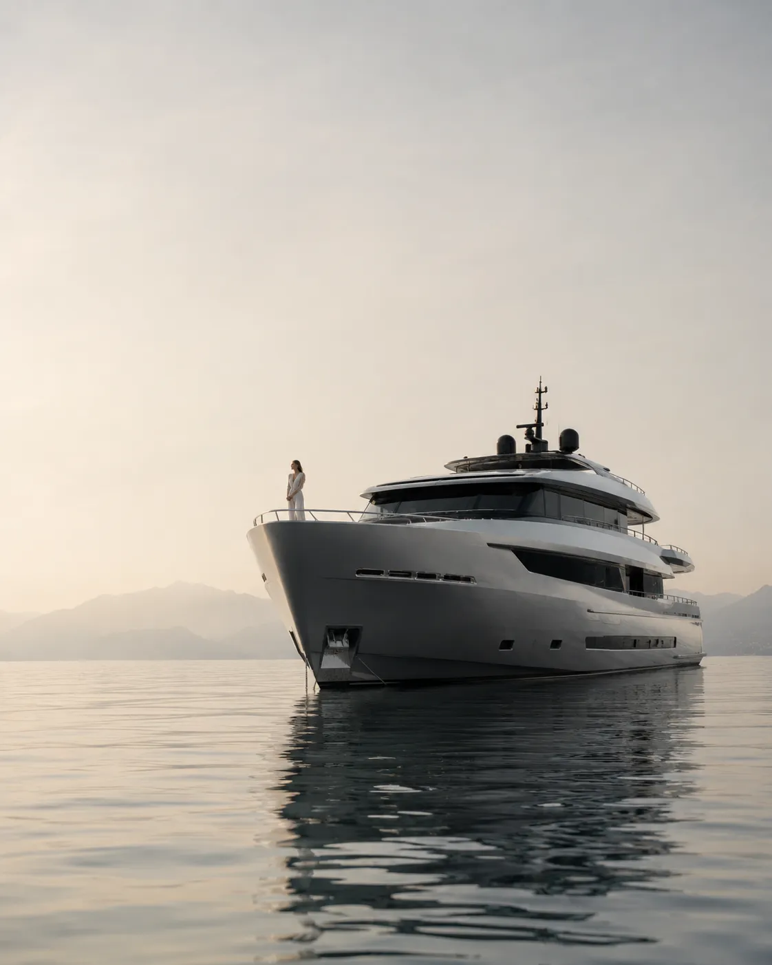 Woman in white dress standing at the bow of a large yacht on calm water with mountains in the background.