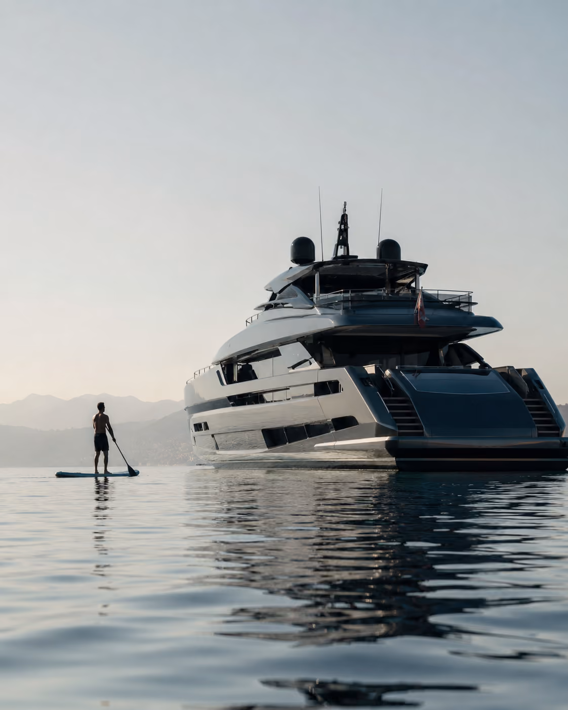 Person paddleboarding near a large yacht on calm water with mountains in the background.