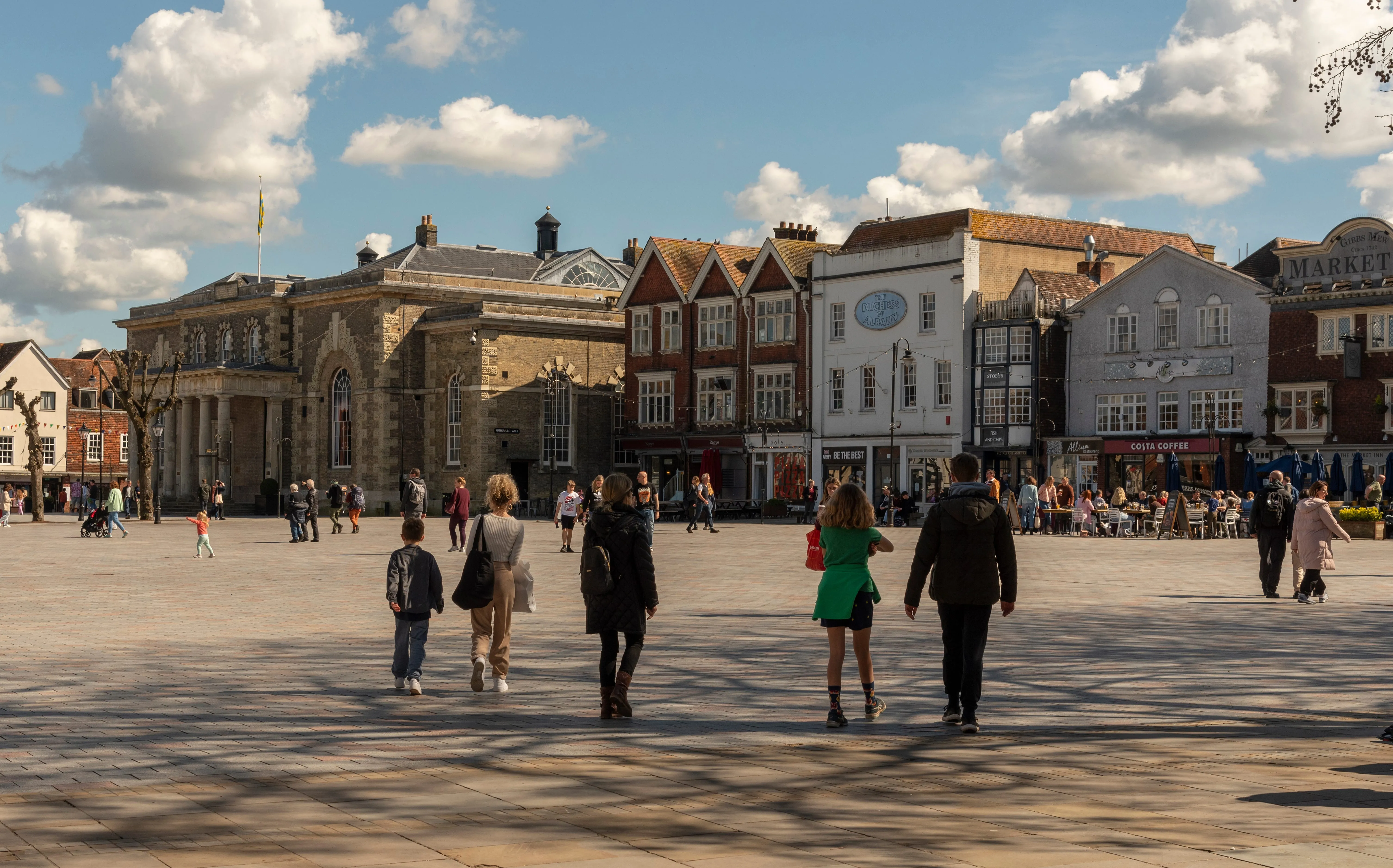 Busy town square with historic stone buildings, colorful facades, and people walking under blue sky