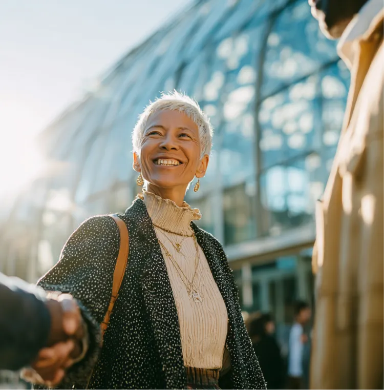 Older woman with short white hair smiling and shaking hands outside a modern glass building.