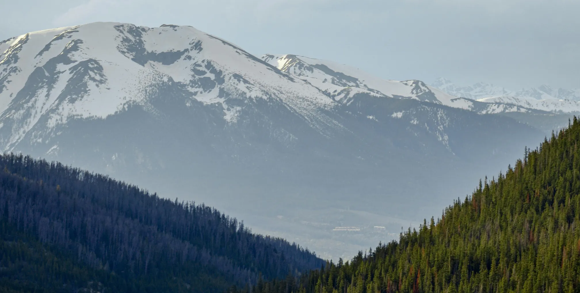Snow-covered mountain peaks behind forested hills under a cloudy sky.