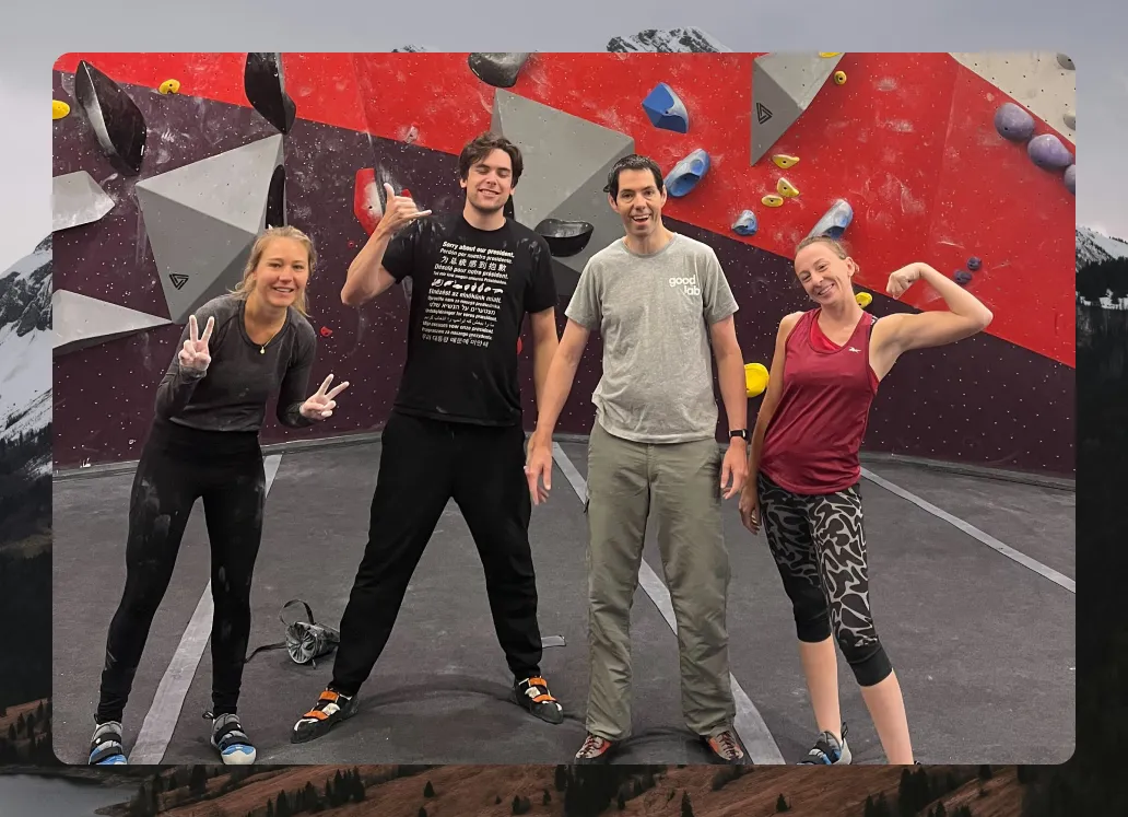 Four people posing and smiling at an indoor climbing gym with colorful climbing holds on walls.