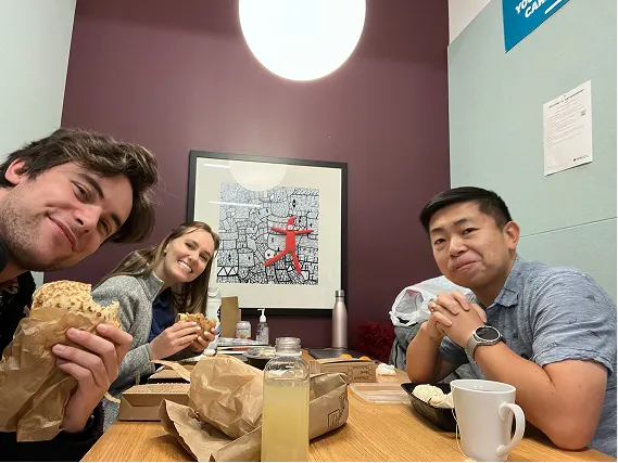 Three people sitting at a table eating sandwiches and food, smiling at the camera in a dining room.