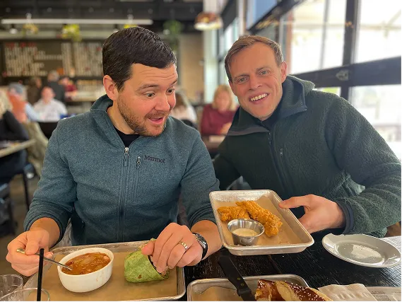 Two men smiling in a restaurant, one holding a tray with fried food, the other eating a wrap and soup.