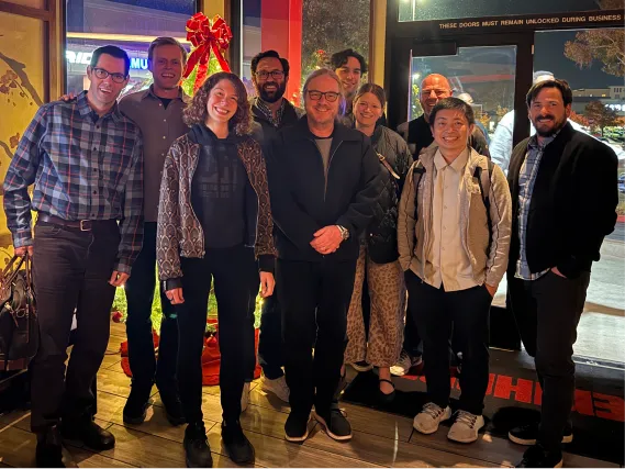 Group of nine diverse people smiling indoors with a Christmas wreath with a red bow in the background.