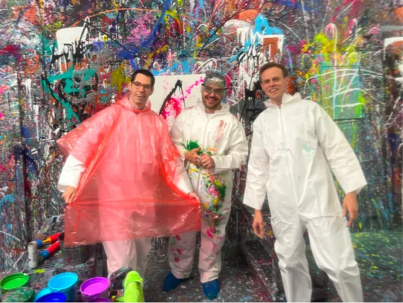 Three men in paint-splattered suits stand in a colorful, messy paint studio with buckets of paint nearby.