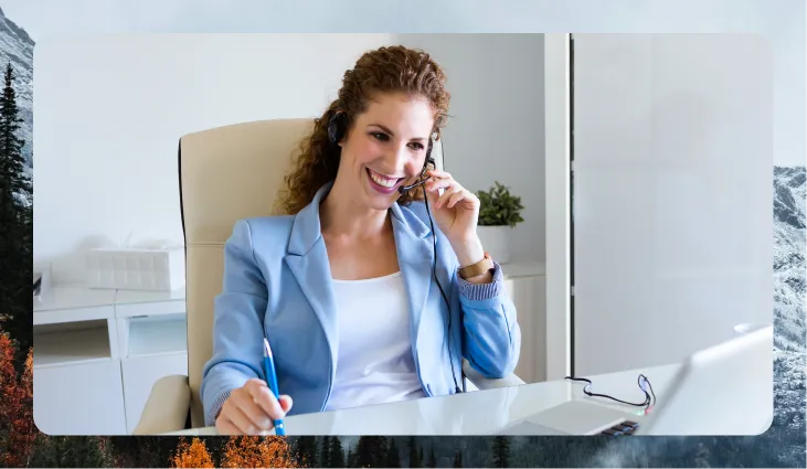 Smiling woman in a blue blazer wearing a headset, holding a pen, and sitting at a desk with a laptop.