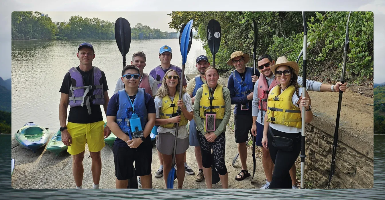 Group of ten people in life jackets holding kayak paddles by a river with kayaks in the background.