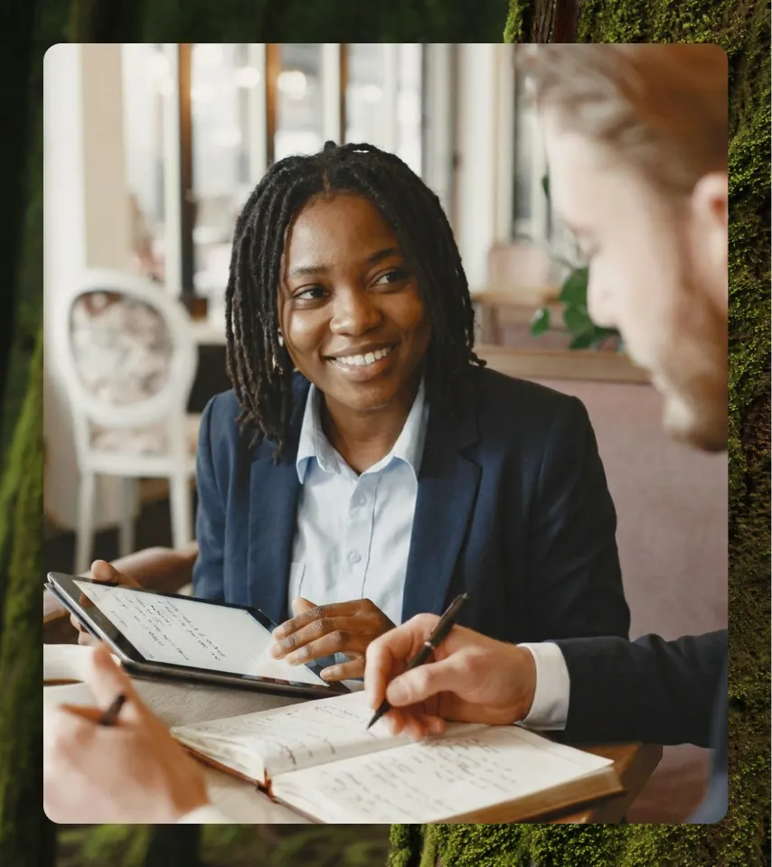 Smiling businesswoman holding a tablet, discussing notes with a colleague writing in a notebook.