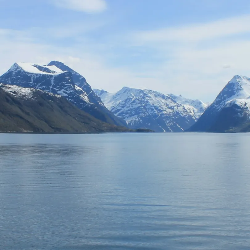 Calm blue fjord surrounded by snow-capped mountains under a partly cloudy sky.