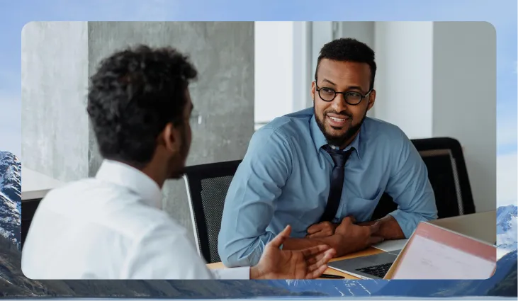 Two men having a discussion at a desk with a laptop in a modern office setting.