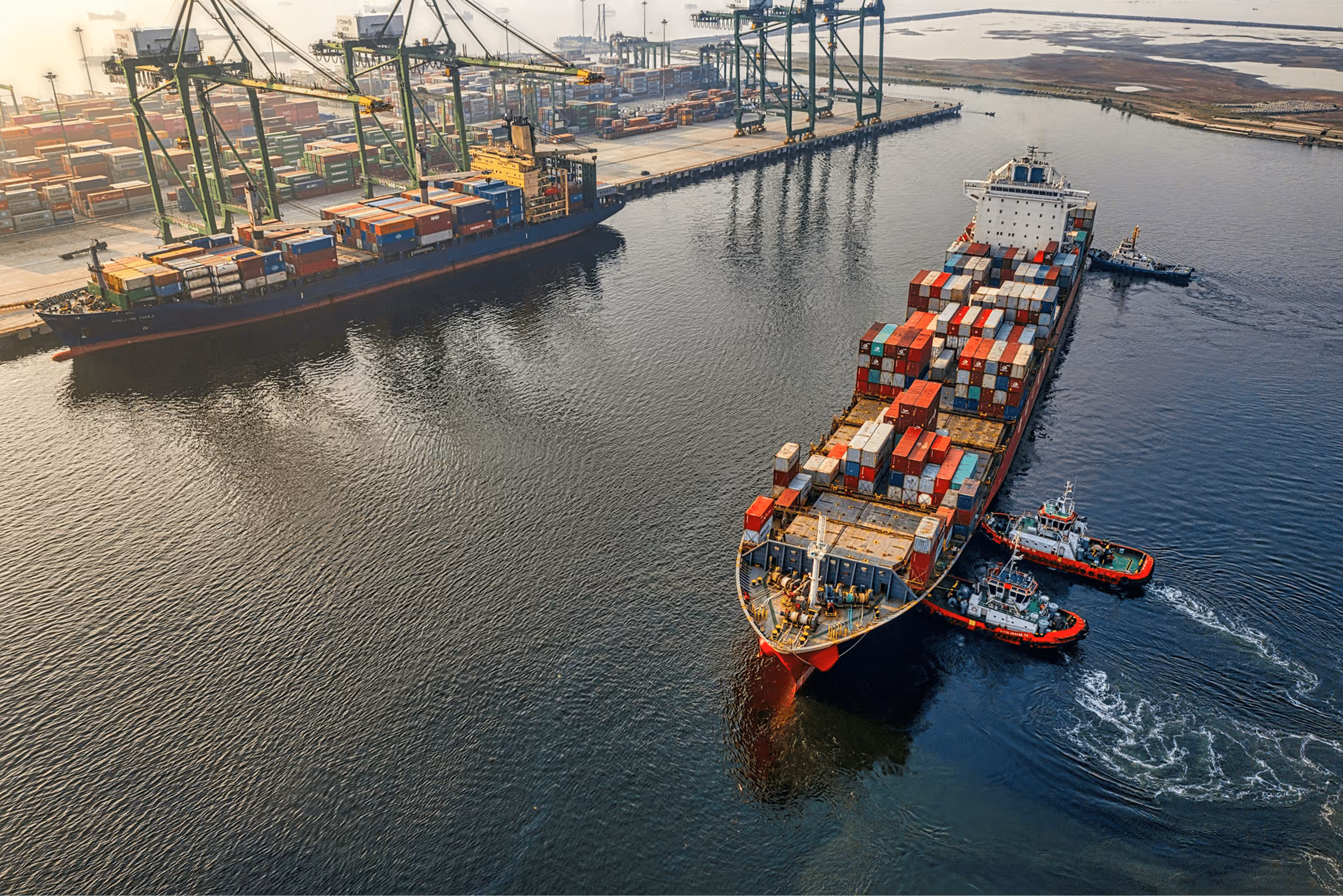 Aerial view of a container ship docked at a harbor with cranes and tugboats in the water.