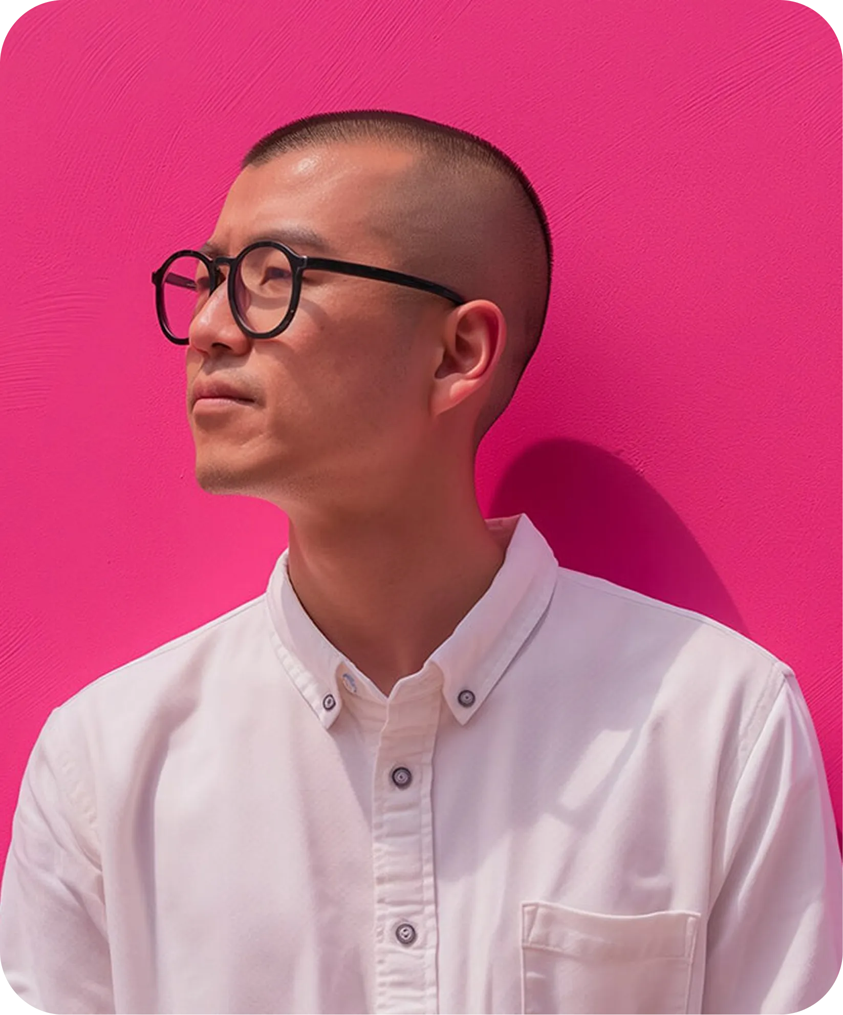 Man with buzz cut and black round glasses wearing a white button-up shirt against a bright pink background.