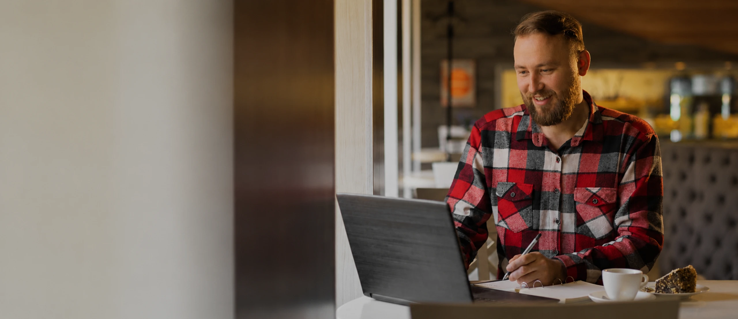 Man in red plaid shirt working on laptop and taking notes with coffee and cake on table.