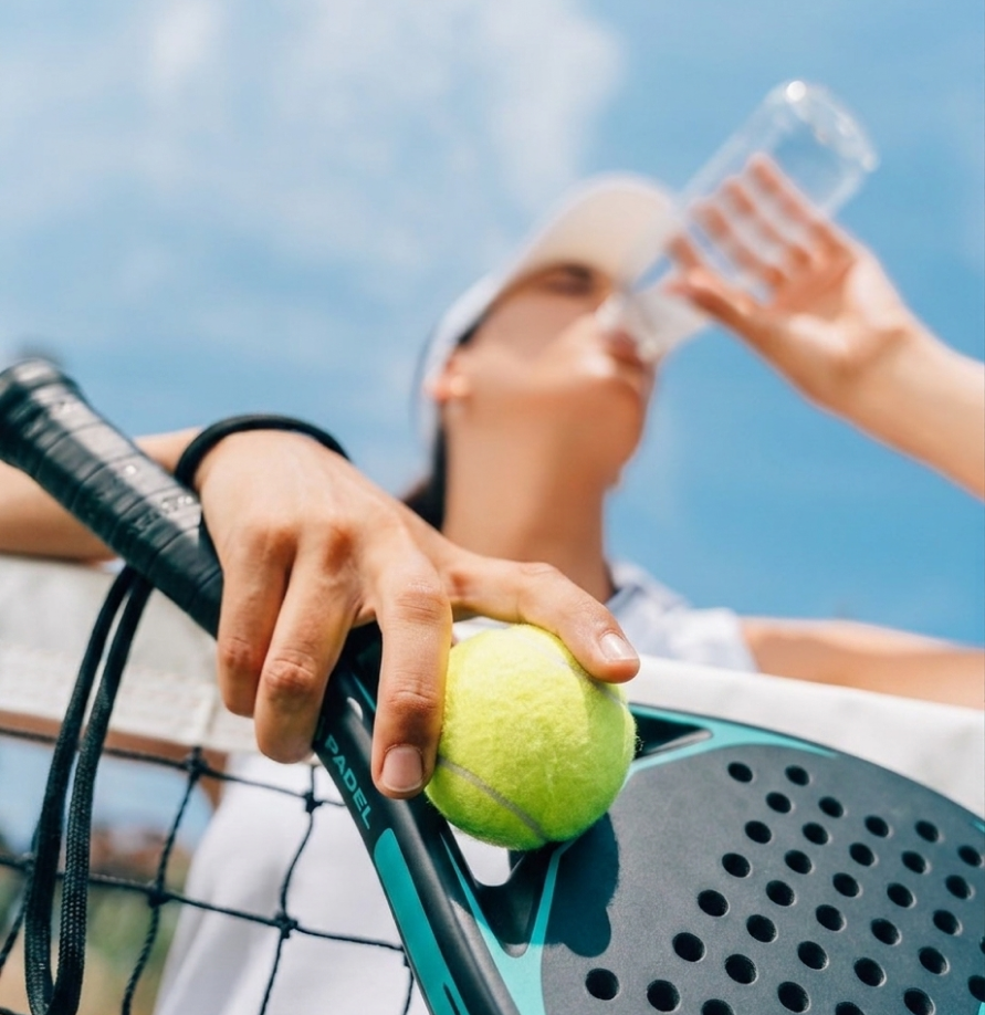 Person holding a padel racket and ball, drinking water with a blue sky background.