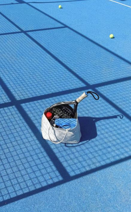 White bag on blue court with a paddle, red ball, and two tennis balls scattered around.