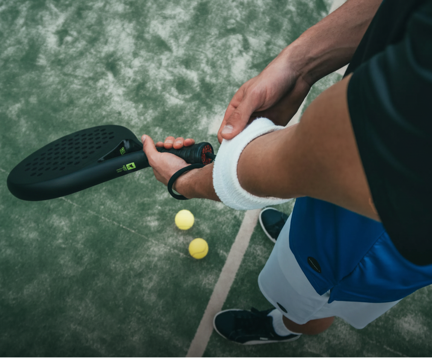 Player on a paddle tennis court holding a black paddle and wearing a white wristband with two balls nearby.