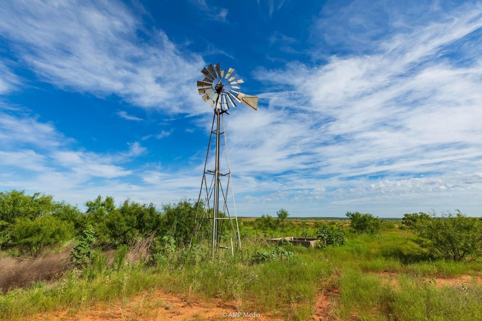Rustic windmill standing in green grassy field under a bright blue sky with scattered clouds.