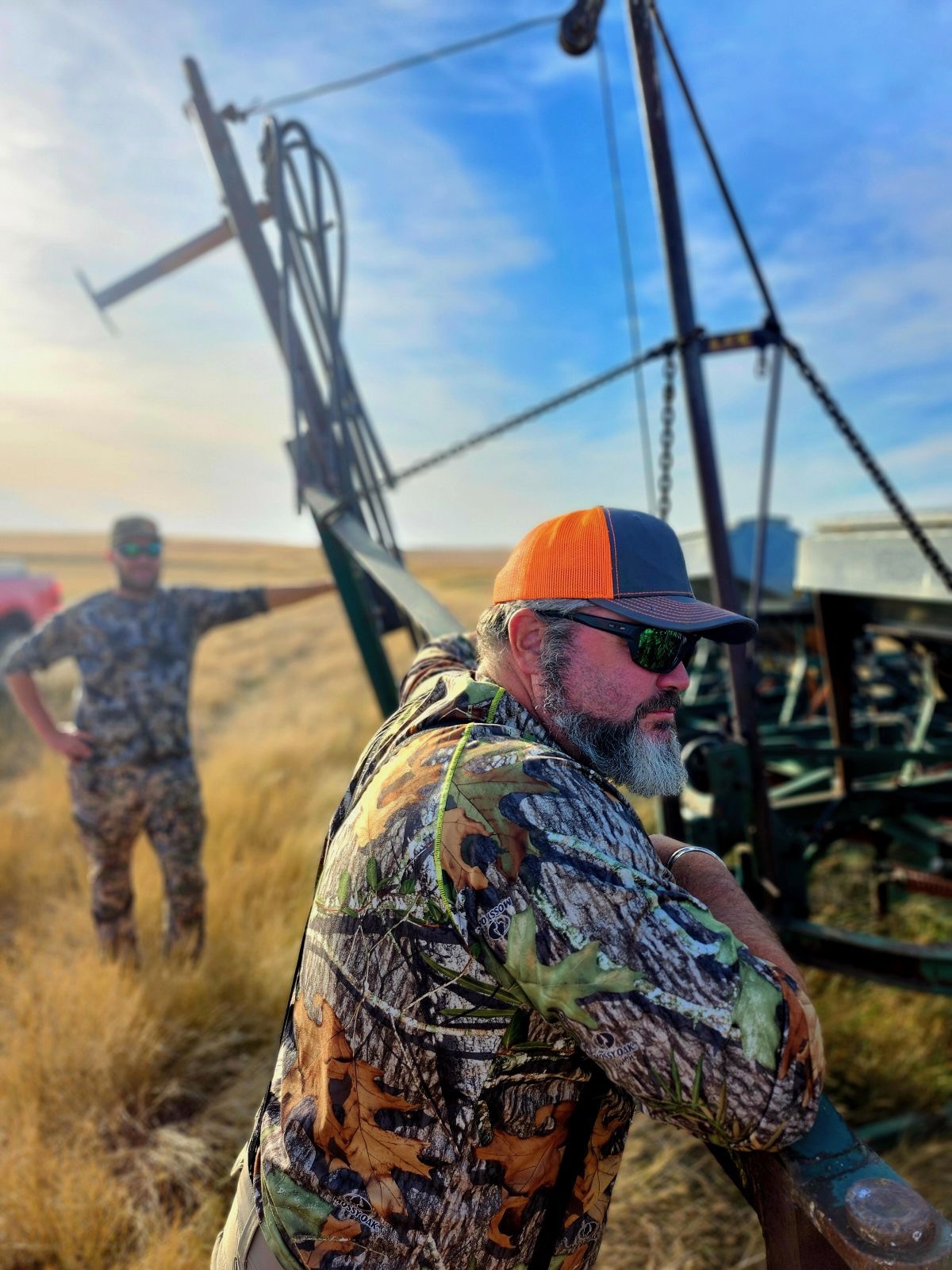 LeRoy Bradley of Wolf Creek Bird Dogs in Mossy Oak camo and a blaze orange cap, leaning on an old farm implement in golden Iowa prairie grass