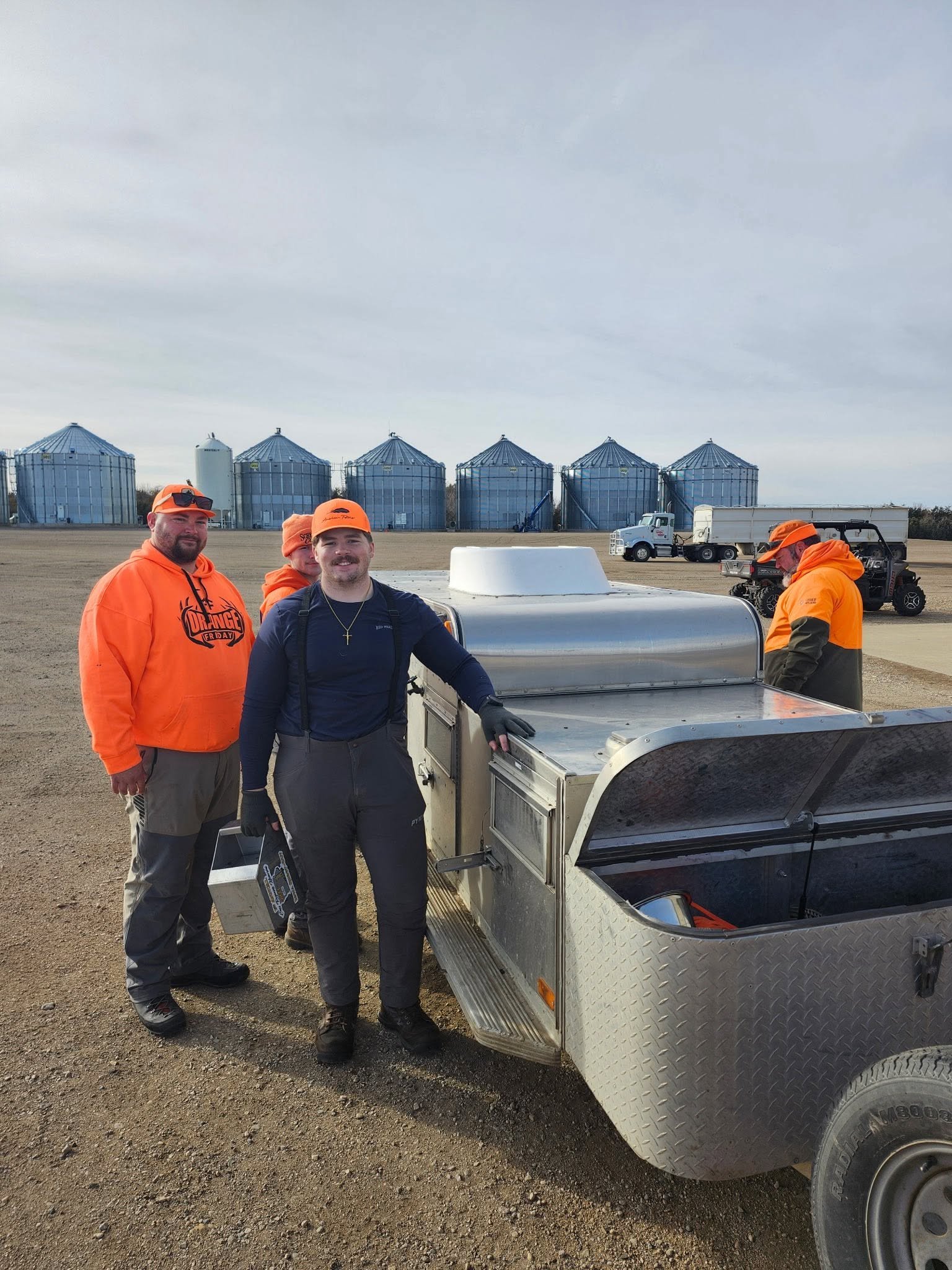 Xander and the crew in blaze orange at the dog trailer before an upland hunt, grain bins behind