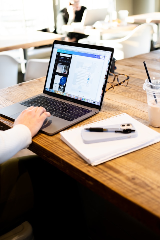 Person working on a laptop at a wooden desk with a notebook, phone, and iced drink beside them