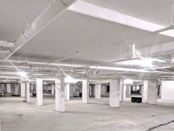 Empty large underground parking garage with white pillars and ceiling pipes under bright lights.